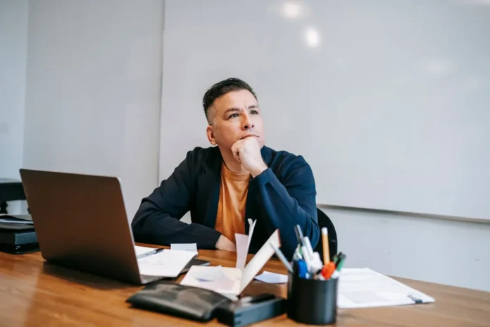 Personne assise à son bureau, regardant par la fenêtre, entourée de documents, dans une posture réfléchie. Personne assise à son bureau, regardant par la fenêtre, entourée de documents, dans une posture réfléchie.