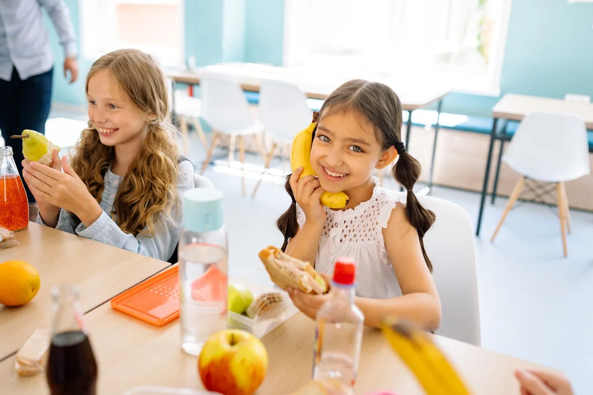 Des enfants mangent à la cantine scolaire, avec des plateaux de nourriture équilibrée devant eux.
